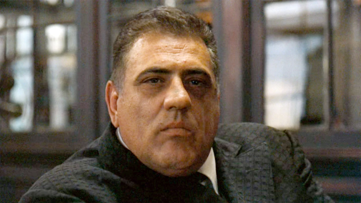 Lenny Montana jako Luca Brasi w filmie "Ojciec chrzestny" Mężczyzna o poważnym wyrazie twarzy ubrany w ciemny garnitur, siedzi na tle ciemnego, eleganckiego wnętrza z dużymi oknami lub szklanymi drzwiami w tle.