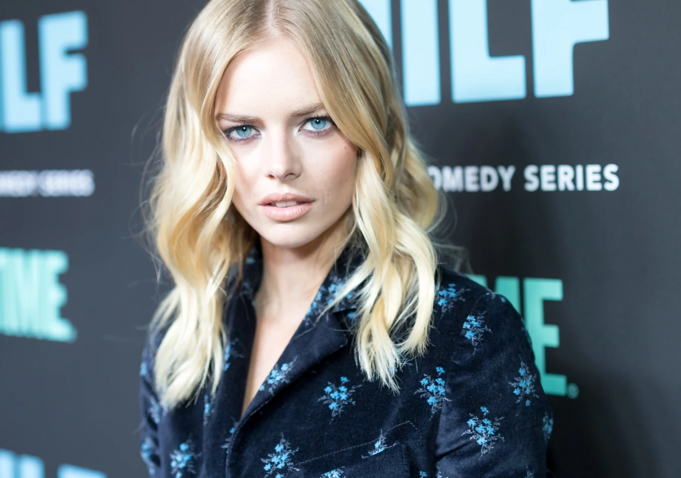 Samara Weaving blondynka o niebieskich oczach w ciemnej marynarce w kwiatowy wzór, z delikatnym makijażem, stoi na tle ściany z napisem i logotypami