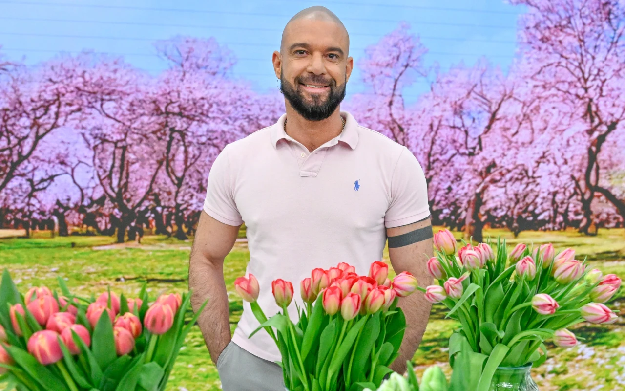 Konrad Gacki Konrad Gacki w różowej koszulce polo stoi za wieloma bukietami różnokolorowych tulipanów w studio "halo tu polsat".