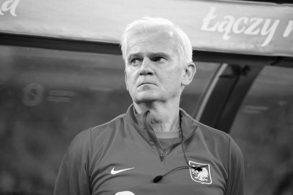Jacek Magiera dojrzały mężczyzna o siwych włosach w sportowej kurtce z godłem Polski i logo Nike, stojący na tle ławki rezerwowych, z poważnym wyrazem twarzy, skoncentrowany na wydarzeniach na boisku