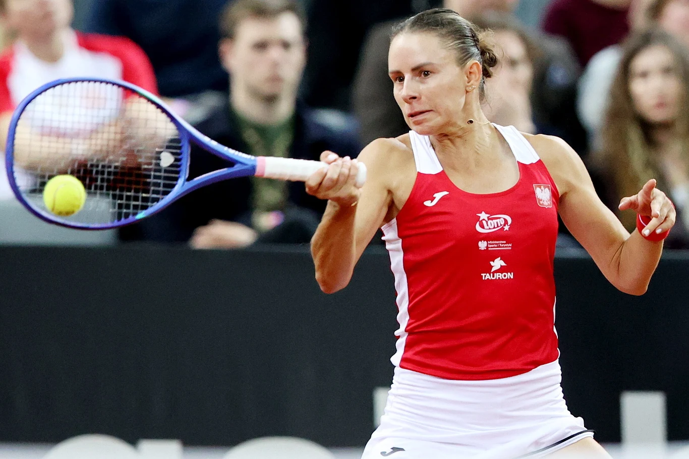 Magda Linette Tenisistka w czerwono-białym stroju wykonuje forehand, skupiona na piłce zbliżającej się do jej rakiety, w tle widoczna publiczność na trybunach.