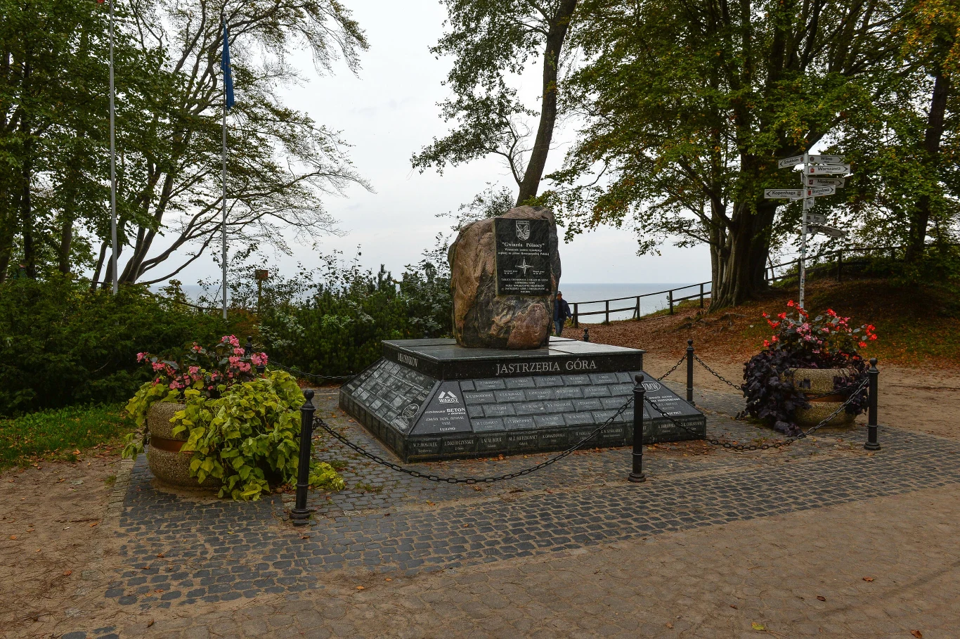 pomnik z czarnego granitu i kamienia otoczony łańcuchem, stojący na brukowanym placu, ozdobiony donicami z kwiatami, wokół drzewa i zielone krzewy, po prawej stronie drewniany drogowskaz