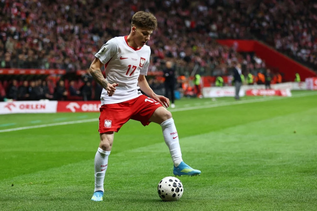 piłkarz reprezentacji Polski w białoczerwonym stroju kontroluje piłkę przy linii bocznej boiska podczas meczu, w tle zapełnione trybuny stadionu
