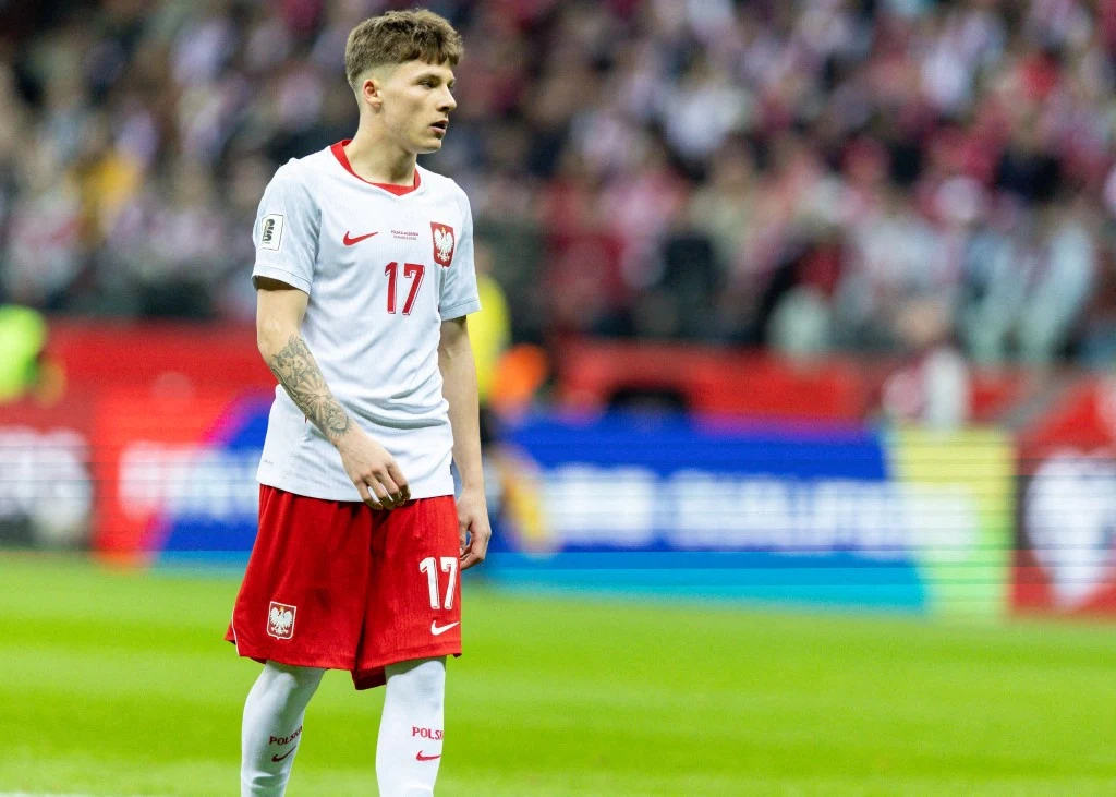 piłkarz w białej koszulce z numerem 17 i czerwonych spodenkach stoi na murawie stadionu podczas meczu, w tle rozmyci kibice na trybunach
