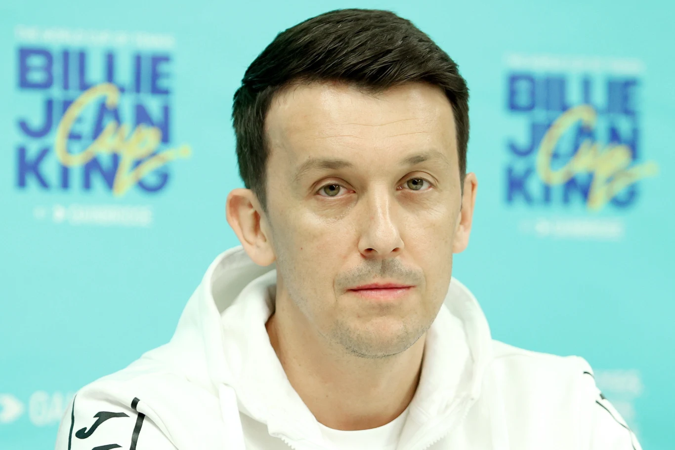 Mężczyzna o krótkich, ciemnych włosach w białej bluzie siedzi na tle niebieskiej planszy z widocznym napisem 'BILLIE JEAN KING'.