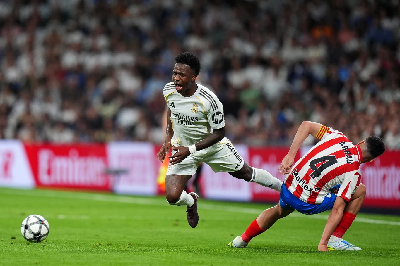 Kadr z meczu Real - Girona piłkarz Realu Madryt w białym stroju ucieka rywalowi z Atletico Madryt, który próbuje go powstrzymać i traci równowagę, dynamiczna akcja rozgrywa się na tle stadionu pełnego kibiców