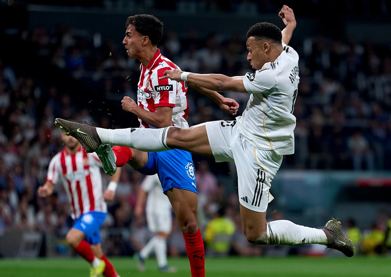 Kadr z meczu Real - Girona Zawodnik Realu Madryt w białym stroju wykonuje wysoki kop przy piłkarzu Girony w czerwono-białych barwach.