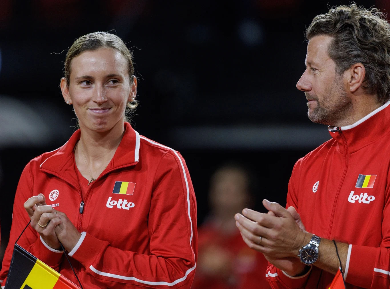 Dwie osoby ubrane w czerwone sportowe bluzy z emblematem belgijskiej flagi i napisem Lotto, stoją obok siebie; kobieta z krótkimi, spiętymi włosami trzyma w rękach małą flagę Belgii i uśmiecha się w stronę mężczyzny, który bije brawo.