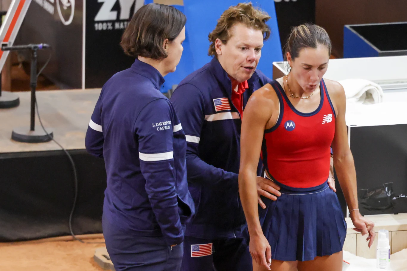 troje członków amerykańskiego zespołu tenisowego podczas przerwy na korcie; kobieta w stroju sportowym z logiem New Balance stoi obok dwóch trenerów w ciemnych dresach z flagą USA, jeden z trenerów wydaje się udzielać wskazówek