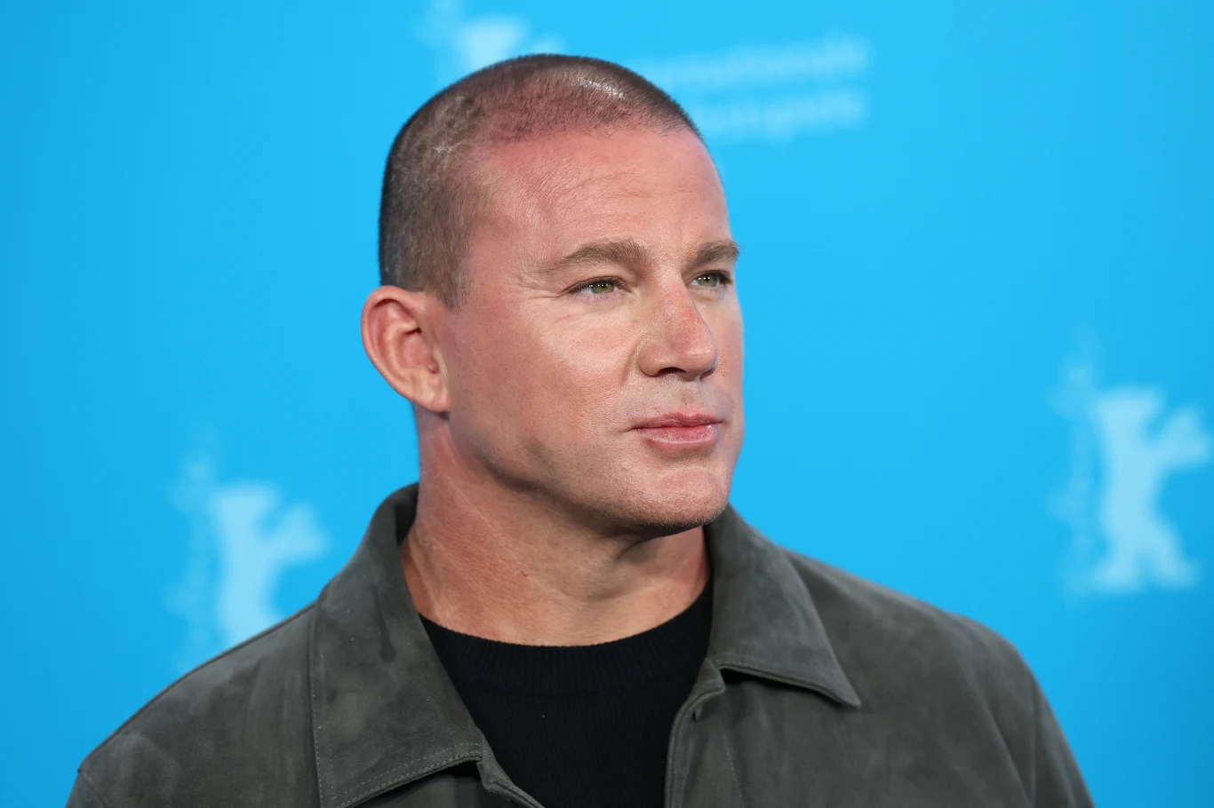 Channing Tatum Mężczyzna o krótkich włosach w szarej kurtce i czarnym podkoszulku stoi na tle niebieskiej ścianki z jasnymi grafikami w tle