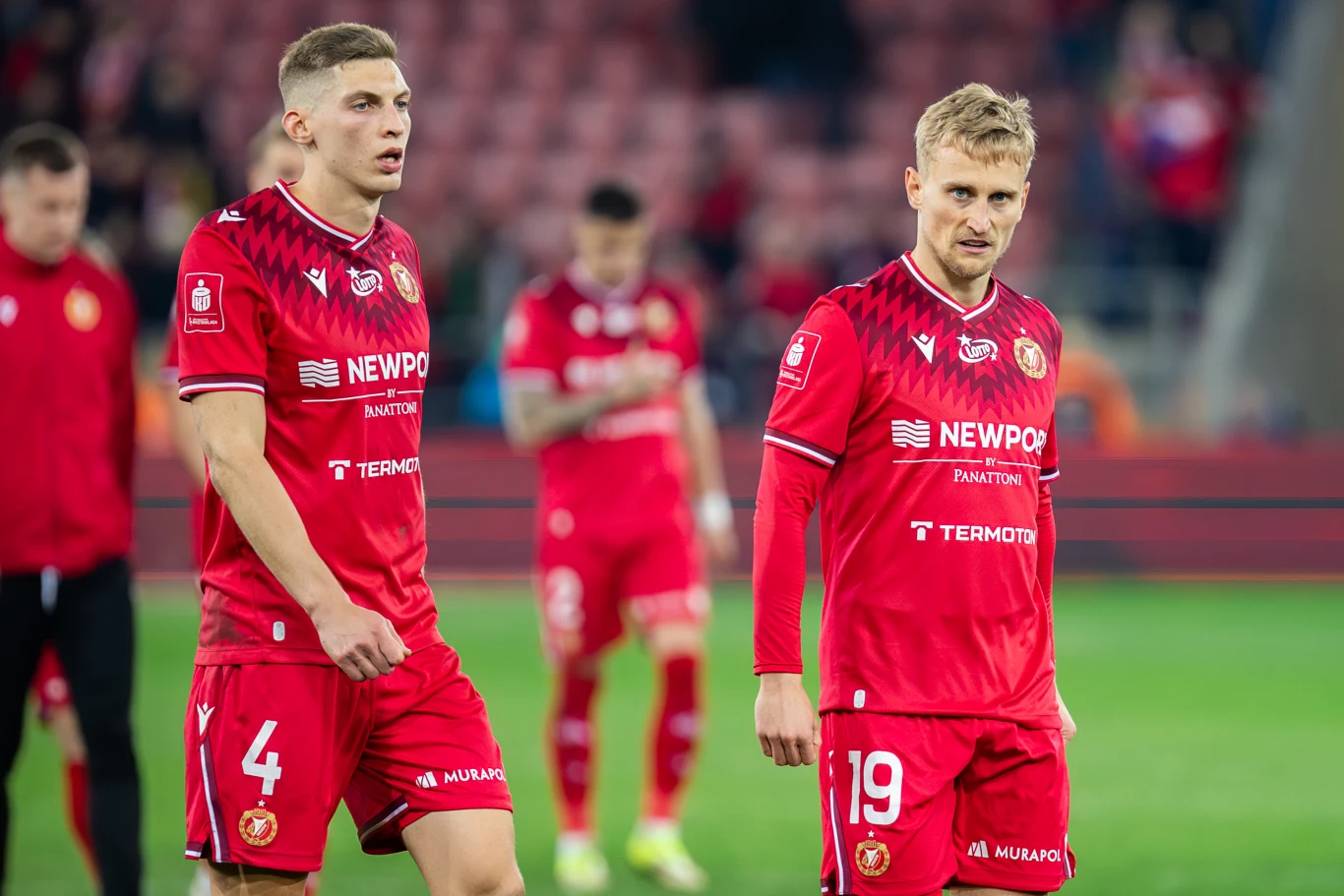 Widzew Łódź znajduje się nad przepaścią Dwóch piłkarzy Widzewa Łódź w czerwonych strojach z numerami 4 i 19 na murawie pustego stadionu.