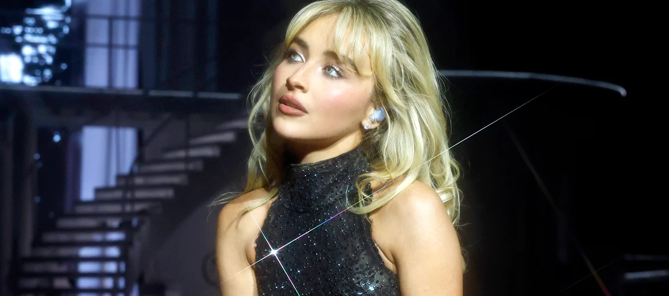 Sabrina Carpenter Sabrina Carpenter w czarnej, błyszczącej sukience na scenie, światło na twarzy, w tle schody.