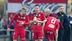 czterech piłkarzy w czerwonych strojach drużyny Widzew Łódź omawia strategię podczas przerwy w grze, jeden z zawodników trzyma bidon, a drugi jest w pochylonej pozycji z numerem 6 na plecach, w tle kibice i transparent z napisem