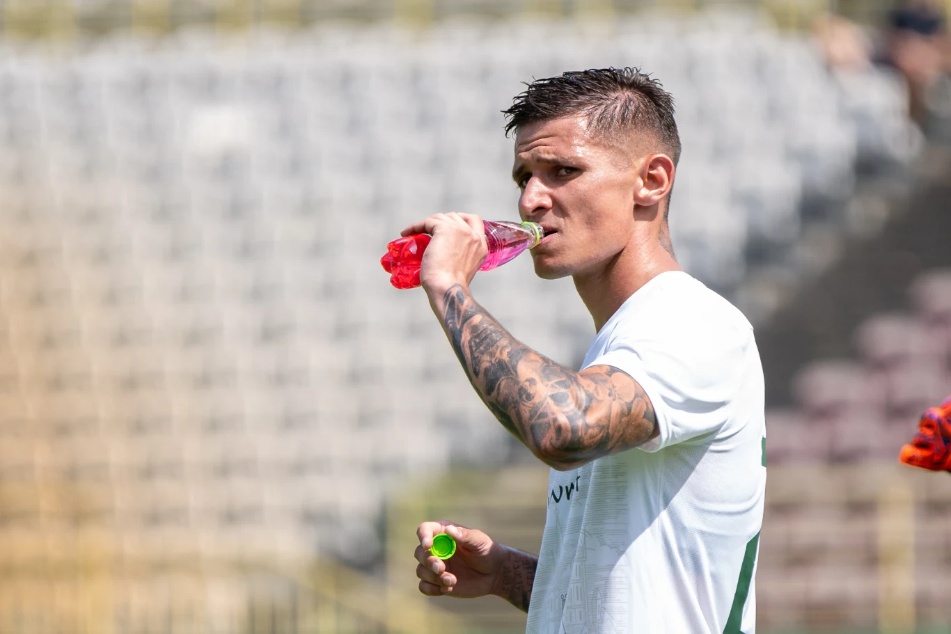 Młody mężczyzna w sportowej koszulce pije różowy napój z butelki, prezentując wytatuowane ramię, za nim rozmyte trybuny stadionu.