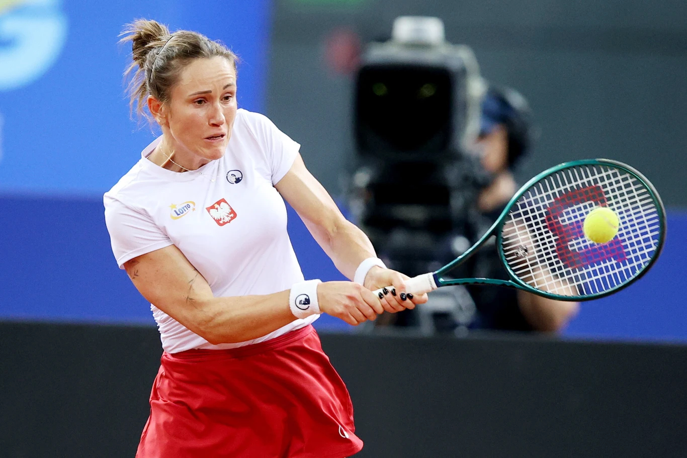 Katarzyna Kawa Tenisistka w białej koszulce z orzełkiem i czerwonej spódniczce wykonuje dynamiczne uderzenie rakietą, koncentrując się na nadlatującej piłce podczas meczu na korcie, w tle widoczna jest kamera telewizyjna.
