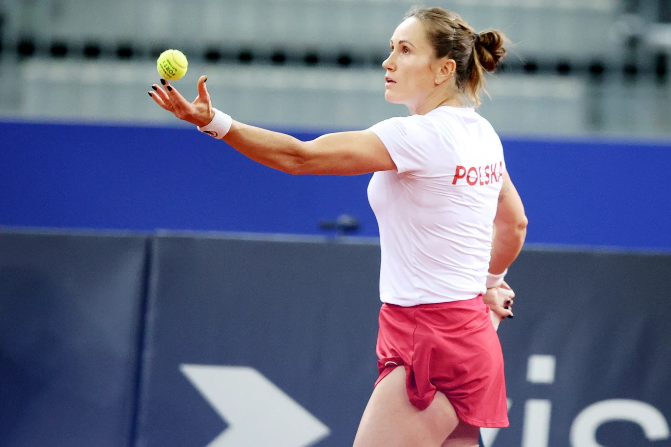 Katarzyna Kawa Tenisistka ubrana w biały t-shirt z napisem 'Polska' i czerwone spodenki koncentruje się przed serwisem, wyrzucając piłkę tenisową do góry podczas meczu na korcie.