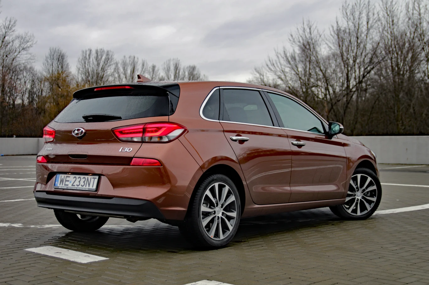 Hyundai i30 hatchback Brązowy Hyundai i30 hatchback zaparkowany na pustym parkingu, widoczny z tyłu na tle bezlistnych drzew i pochmurnego nieba.