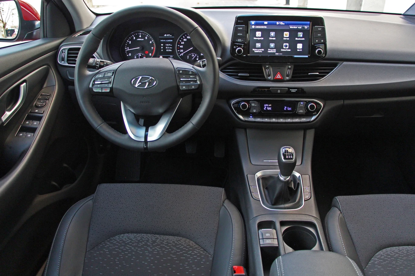 Hyundai i30 kokpit samochodu Hyundai z wyraźnie widocznym wielofunkcyjnym kierownicą, nowoczesnym panelem środkowym z ekranem dotykowym, manualną skrzynią biegów i ergonomicznym rozmieszczeniem przycisków na desce rozdzielczej