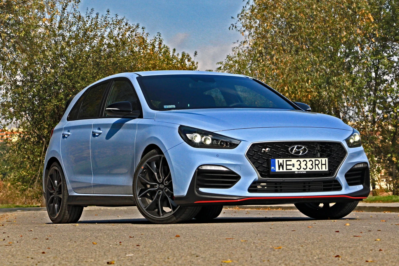jasnoniebieski Hyundai i30 N hatchback na czarnych felgach stoi na asfalcie, na tle drzew i nieba