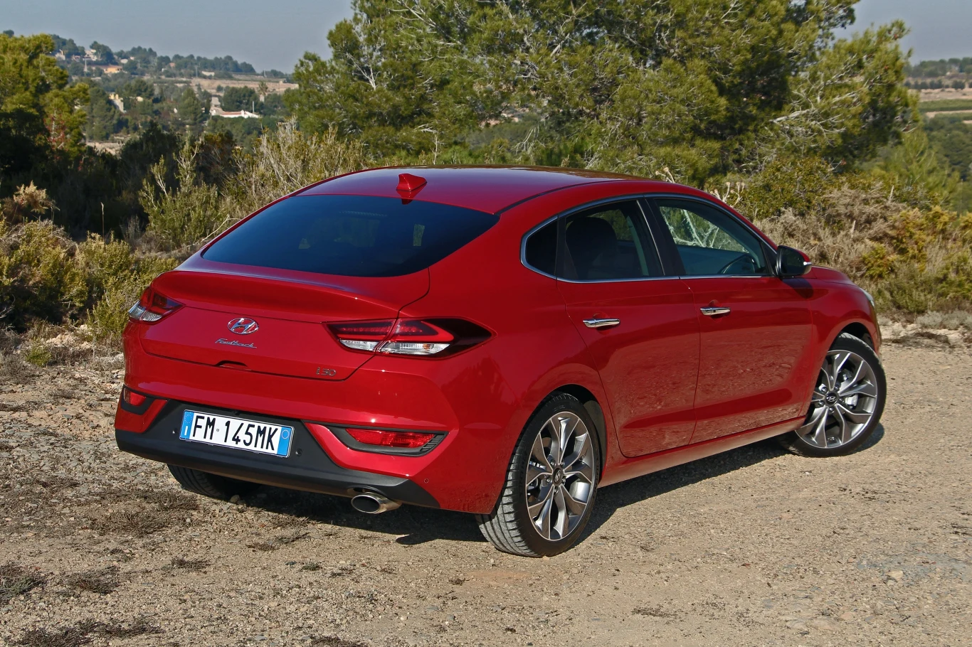 Hyundai i30 Fastback czerwony samochód Hyundai Elantra w sportowej wersji, zaparkowany na szutrowym podłożu, otoczony zielonymi krzewami i drzewami, w tle widoczne wzgórza oraz jasne niebo