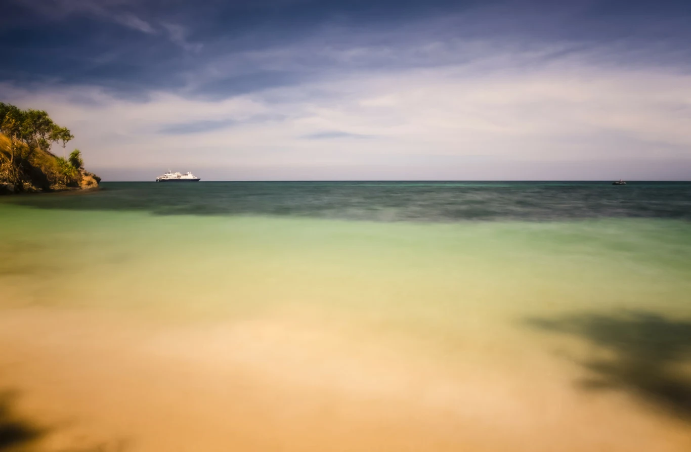 Spokojna, piaszczysta plaża z przejrzystą wodą w odcieniach turkusu, na horyzoncie widoczny duży biały statek, po lewej stronie fragment bujnej zieleni nadbrzeżnych drzew.
