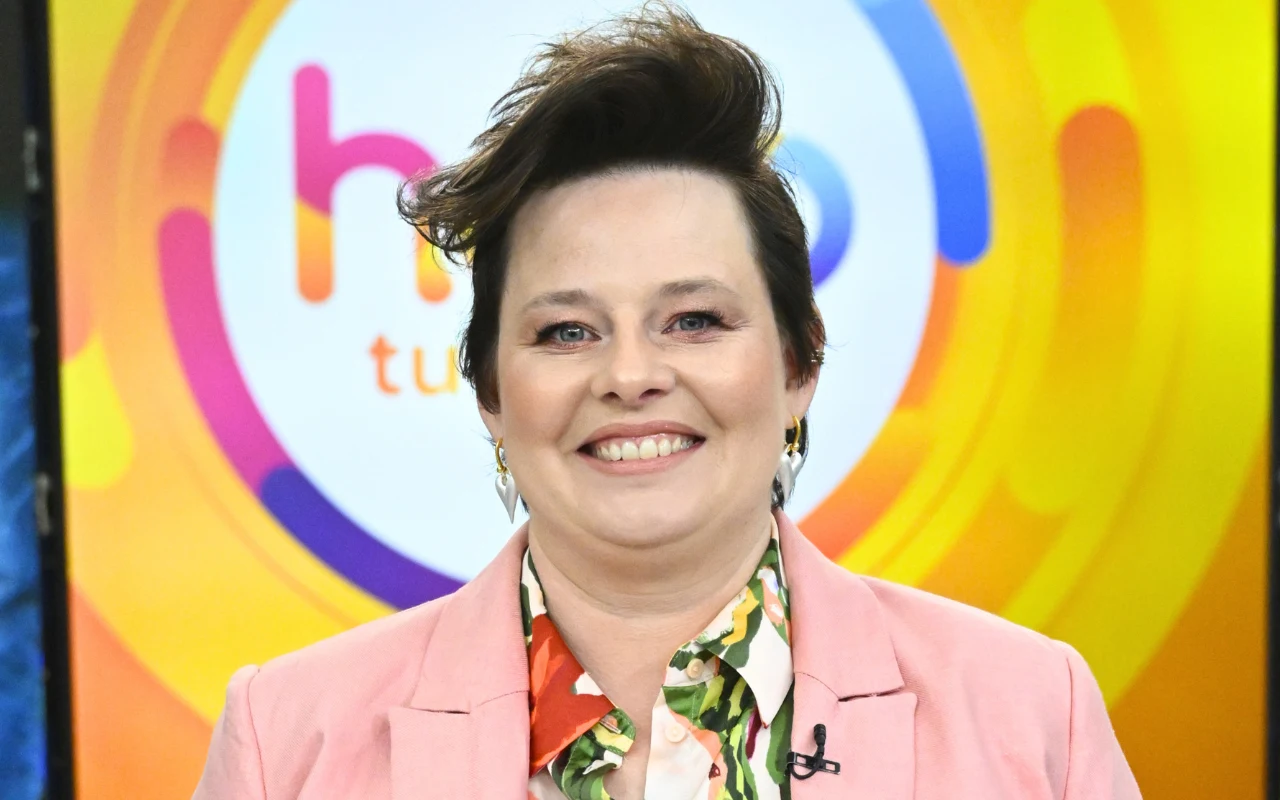 Małgorzata po metamorfozie prezentowała się zachwycająco Uśmiechnięta kobieta w różowej marynarce oraz kolorowej koszuli, stojąca na tle kolorowego logo programu telewizyjnego z dominującymi odcieniami żółci, różu i niebieskiego.