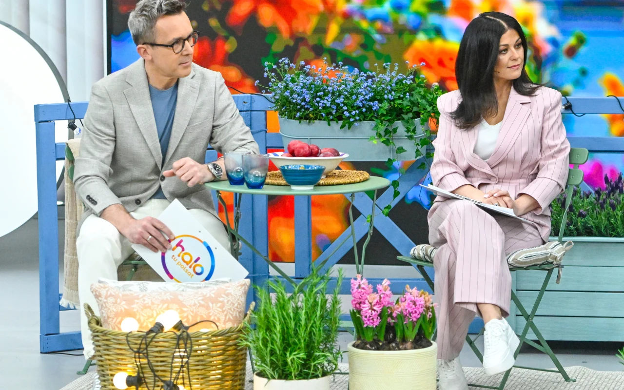 Prowadzący "halo tu polsat" Dwójka prowadzących siedzi na kolorowo urządzonej scenie, otoczonej kwiatami i dekoracjami ogrodowymi; pomiędzy nimi mały stolik z naczyniami i owocami, w tle wyraziste, kolorowe obrazy kwiatów.