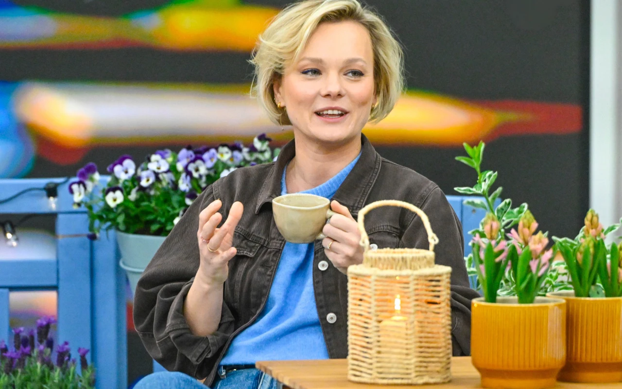 Kasia Joda pokazuje, jak zaaranżować balkon. Uśmiechnięta kobieta z jasnymi włosami siedzi przy drewnianym stole, trzymając w dłoni filiżankę, obok niej stoi pleciony lampion z zapaloną świecą oraz doniczki z kwitnącymi kwiatami, w tle widoczne są skrzynki i kolorowe bratki.
