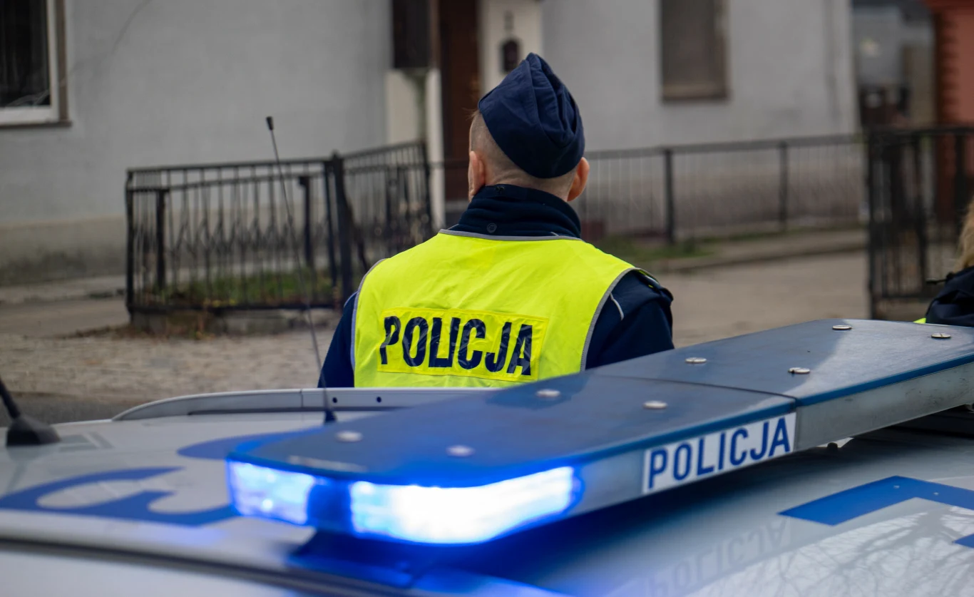 Pod Warszawą znaleziono niezidentyfikowany balon (zdj. ilustracyjne) Policjant w kamizelce 'Policja' przy radiowozie z niebieskim światłem, ogrodzenie i dom w tle.