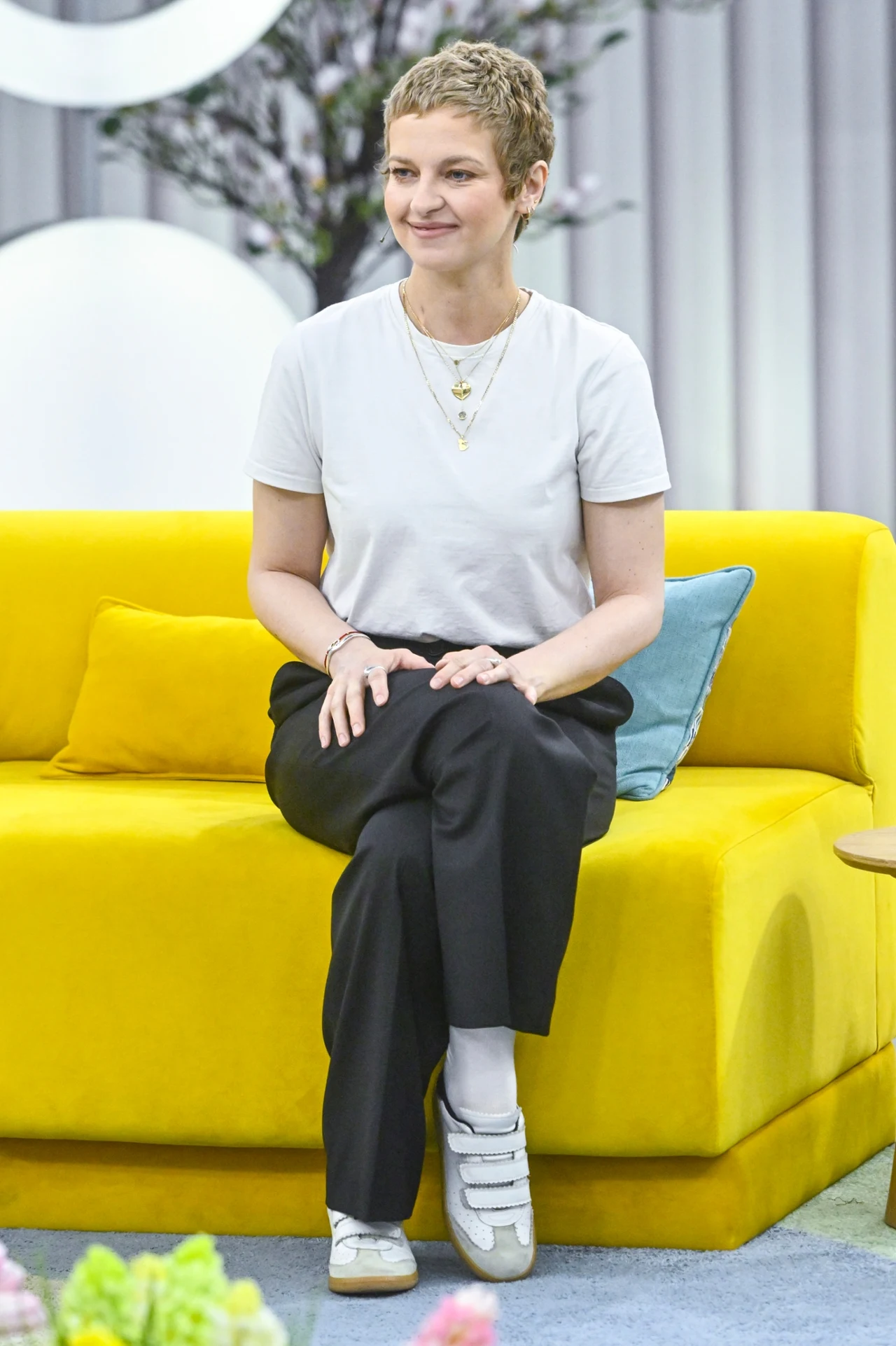 Marta Nieradkiewicz Młoda kobieta z krótkimi włosami siedzi na jasnożółtej sofie, ubrana w białą koszulkę, czarne spodnie i białe sportowe buty, obok niej jasnoniebieska poduszka; w tle delikatna dekoracja z kwiatami i geometrycznymi elementami.