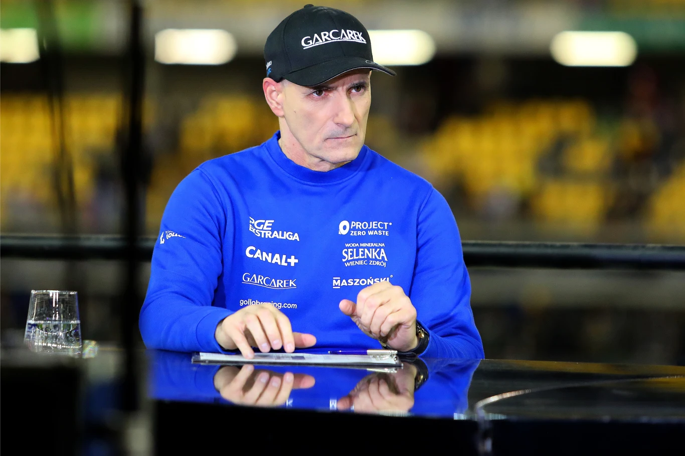 Tomasz Gollob Tomasz Gollob w niebieskiej bluzie z logotypami, czapce, przy stole z tabletem i wodą; w tle żółte krzesła stadionowe