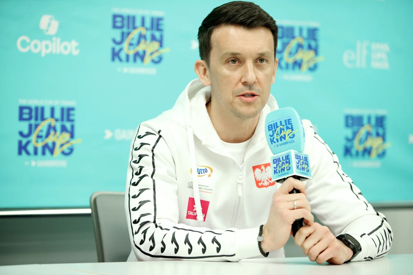 mężczyzna w białej sportowej bluzie z mikrofonem w ręku udziela wywiadu na tle ścianki z logotypami Billie Jean King Cup oraz sponsorów