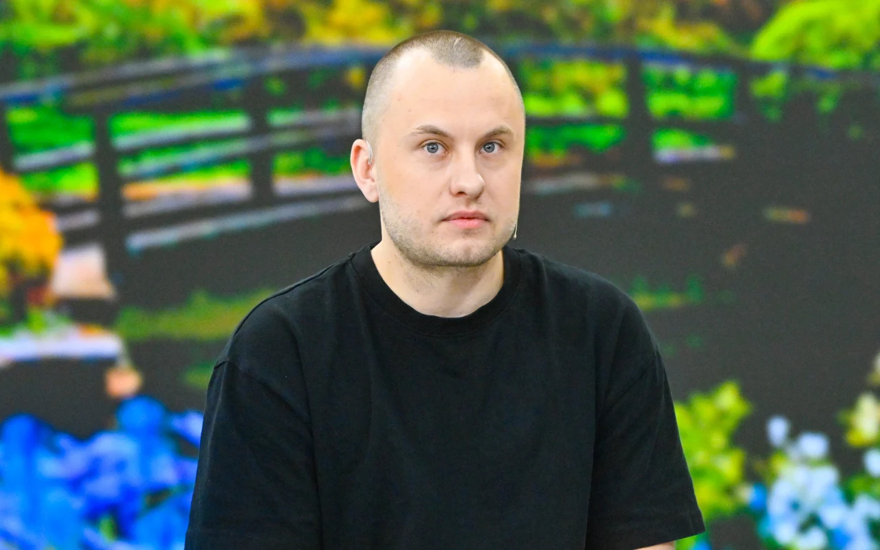 mężczyzna z krótkimi włosami w czarnej koszulce siedzi na tle kolorowej tablicy z motywem ogrodu i mostku