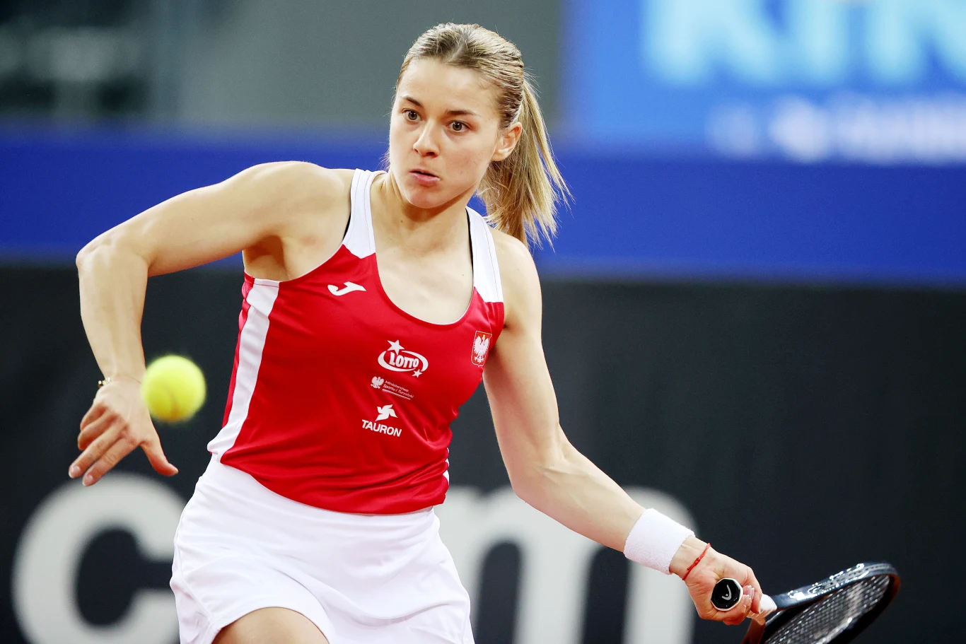 Zawodniczka w czerwono-białym stroju sportowym podczas dynamicznej akcji tenisowej, skoncentrowana na odbiciu żółtej piłki rakietą.