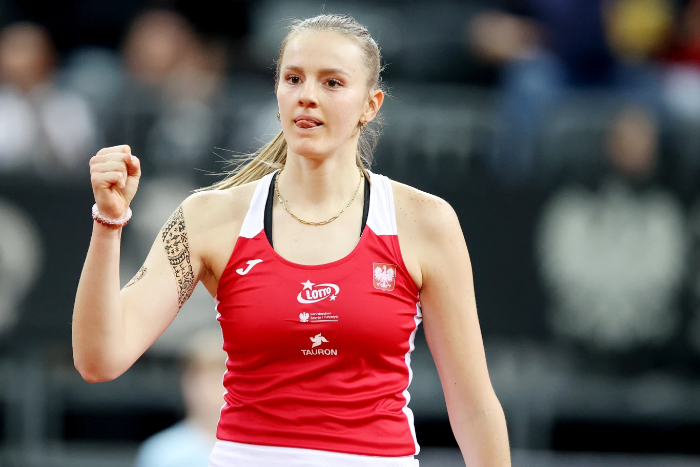 Młoda kobieta w sportowej koszulce z polskim godłem i logotypami sponsorów, unosi zaciśniętą pięść w geście triumfu po udanej akcji, skupiona i pewna siebie na tle rozmytej publiczności.