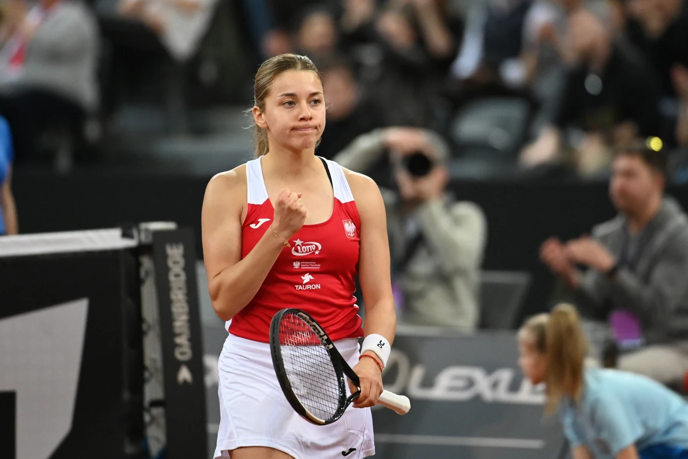 Maja Chwalińska tenisistka w czerwono-białym stroju sportowym z rakietą w dłoni, ściskająca pięść w geście triumfu na korcie, w tle rozmyta publiczność