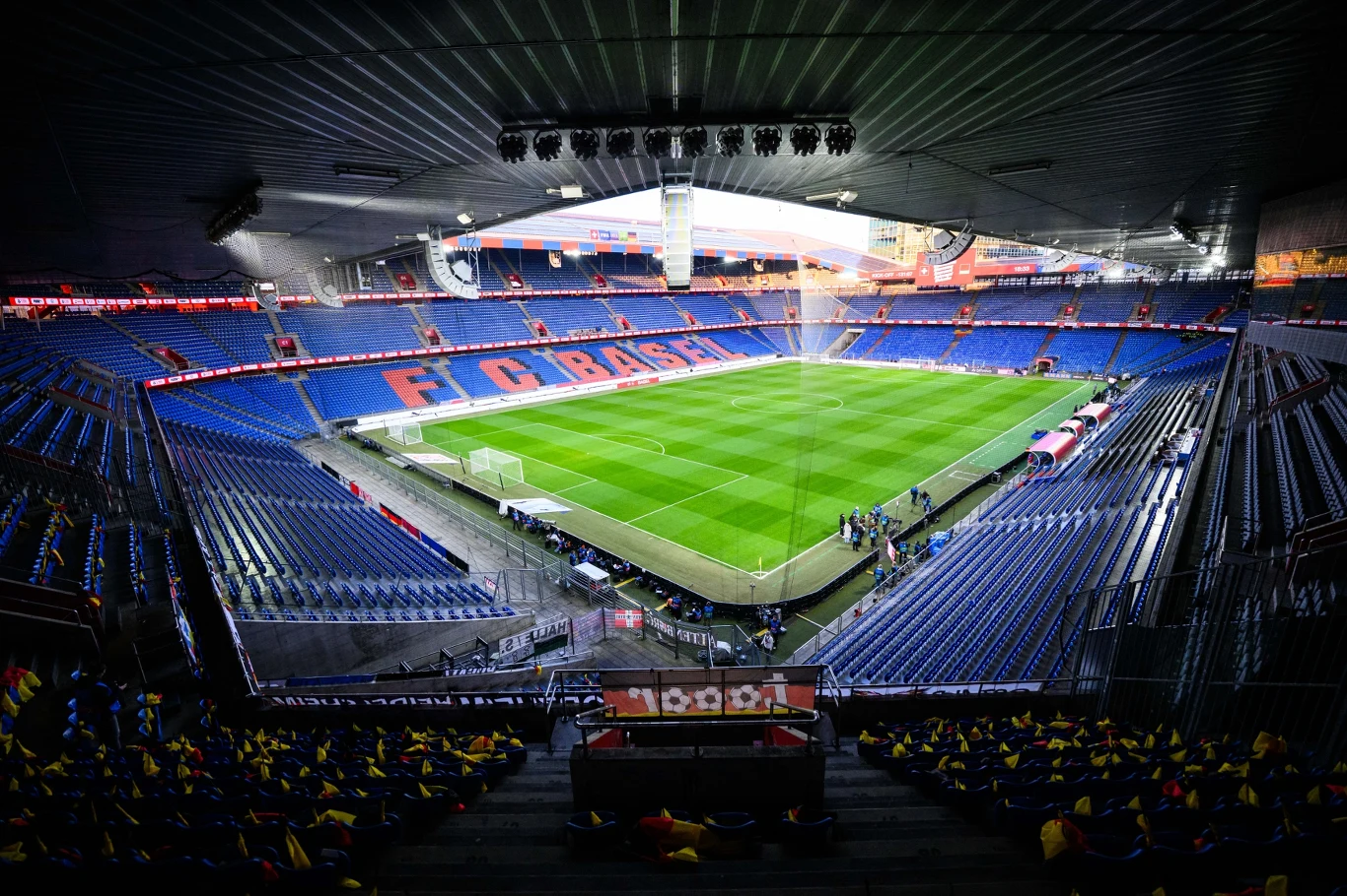 nowoczesny stadion piłkarski z niebieskimi i czerwonymi trybunami, na których widoczny jest napis 'F.C. Basel', zielona murawa boiska oraz kilka osób przygotowujących pole gry