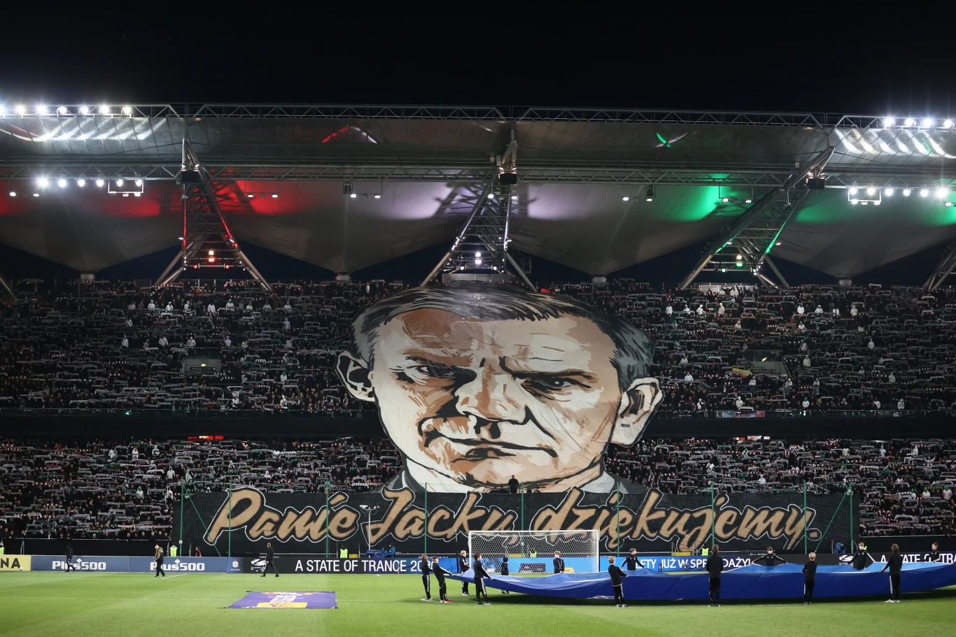 Ogromna sektorówka z wizerunkiem mężczyzny dominuje trybuny piłkarskiego stadionu, poniżej widoczny jest napis „Panie Jacku dziękujemy”, a kibice trzymają szaliki nad głowami w geście uznania, podkreślając atmosferę hołdu i wdzięczności.