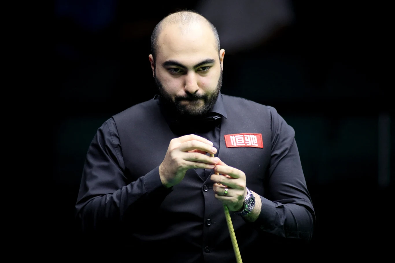 Hossein Vafaei mężczyzna o łysiejących włosach w ciemnym stroju bilardowym trzyma kij snookerowy, skupiony na przygotowaniach do gry, oświetlony światłem na tle ciemnego, rozmytego tła