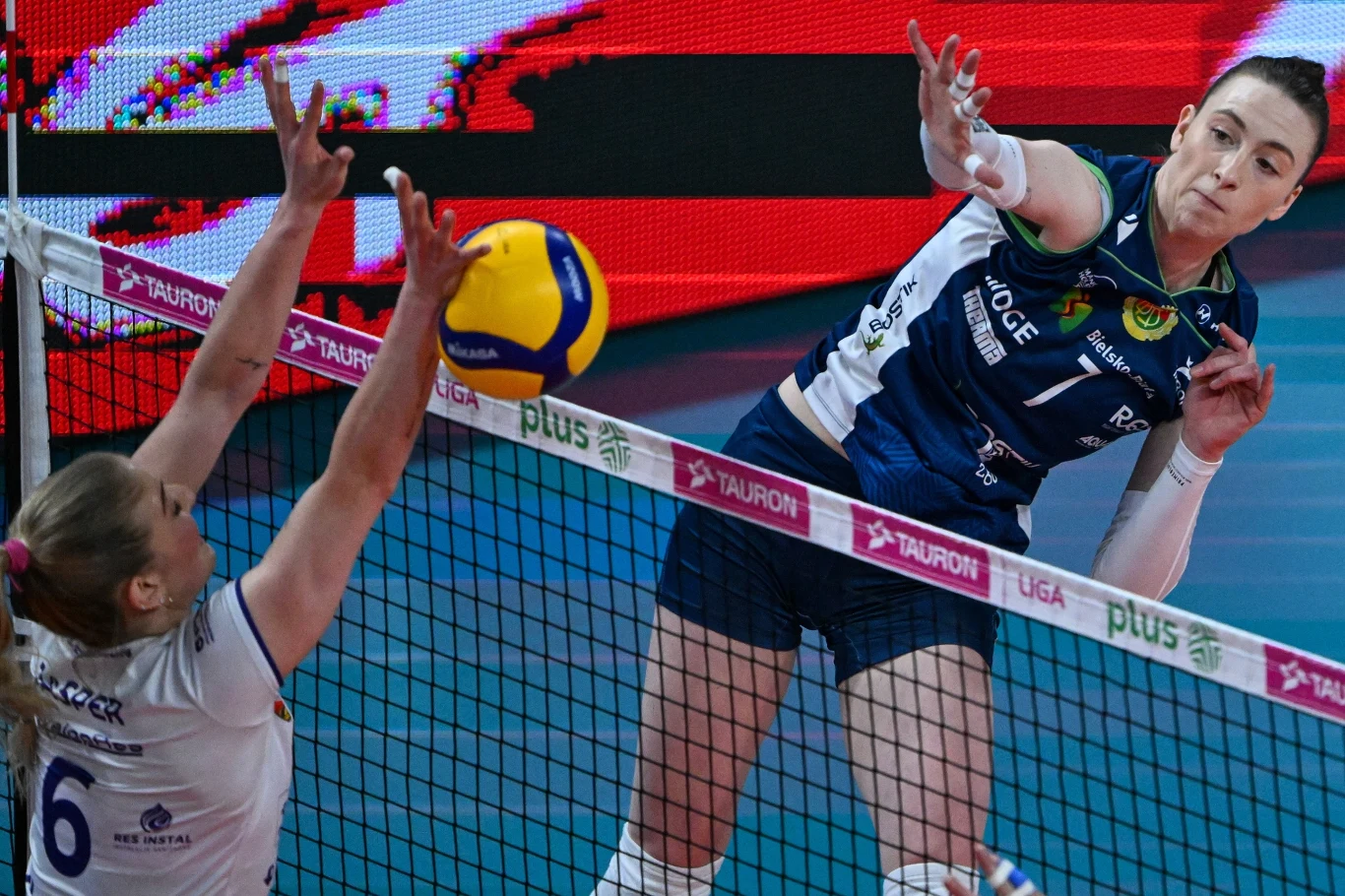 Zawodniczka w granatowej koszulce atakuje piłkę siatkową nad siatką, próbując zdobyć punkt, podczas gdy przeciwniczka w białym stroju próbuje ją zablokować, obie wykazując pełne zaangażowanie w dynamicznej akcji meczowej.