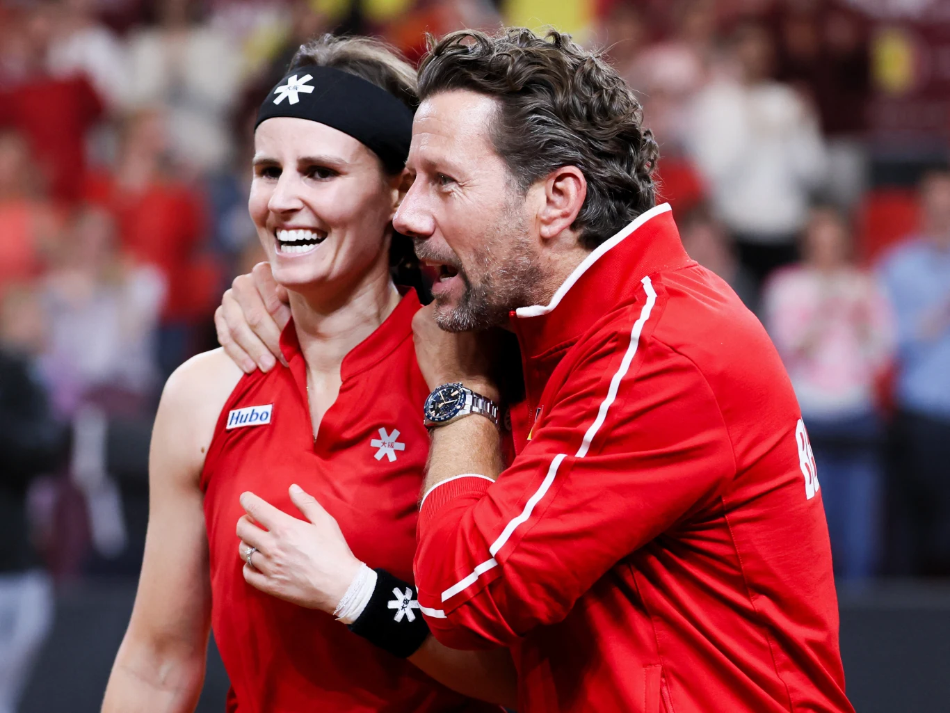 Wim Fissette i Greet Minen dwoje uśmiechniętych ludzi ubranych w sportowe czerwone stroje, kobieta z opaską na głowie i mężczyzna przytulający ją ramię w ramię, w tle rozmyta publiczność