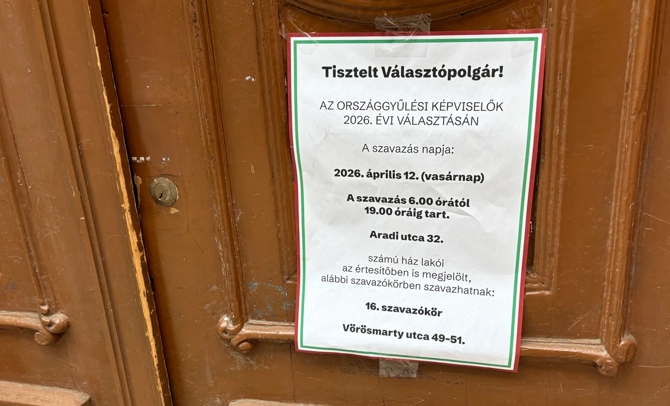 Oficjalna informacja wyborcza w języku węgierskim, wydrukowana na białej kartce i przyklejona na brązowych drzwiach, zawiadamiająca o terminie i szczegółach głosowania do parlamentu w 2026 roku, ze szczegółową lokalizacją i godzinami otwarcia lokalu wy...