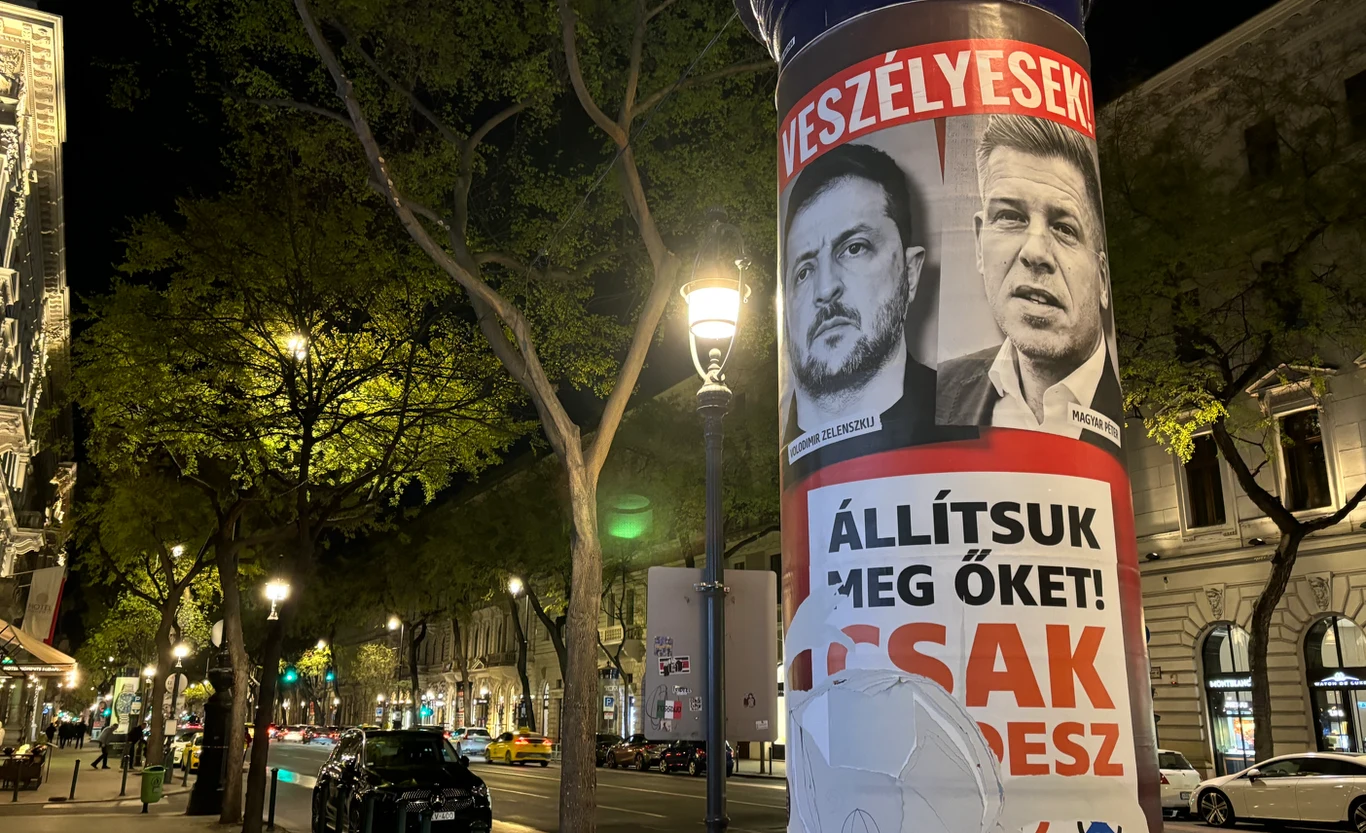 "Są niebezpieczni". Plakat kampanii Fideszu plakat wyborczy z dużymi portretami dwóch mężczyzn i hasłami w języku węgierskim przyklejony na słupie ogłoszeniowym, ustawionym przy oświetlonej ulicy z wysokimi drzewami i historycznymi kamienicami w tle