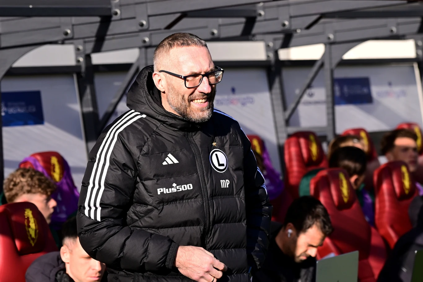 Mężczyzna w okularach ubrany w czarną kurtkę sportową z logo Legii Warszawa stoi przed ławką rezerwowych na stadionie, w tle kilku mężczyzn również w sportowych ubraniach siedzi na fotelach z burgundowymi oparciami i żółtym emblematem.