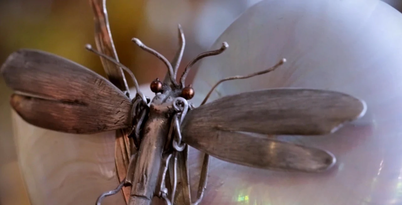 Ważka z warsztatu jubilerskiego Jana Kalińskiego metalowy owad o dużych skrzydłach z wypukłymi oczami i cienkimi czułkami przymocowany do błyszczącej, perłowej powierzchni