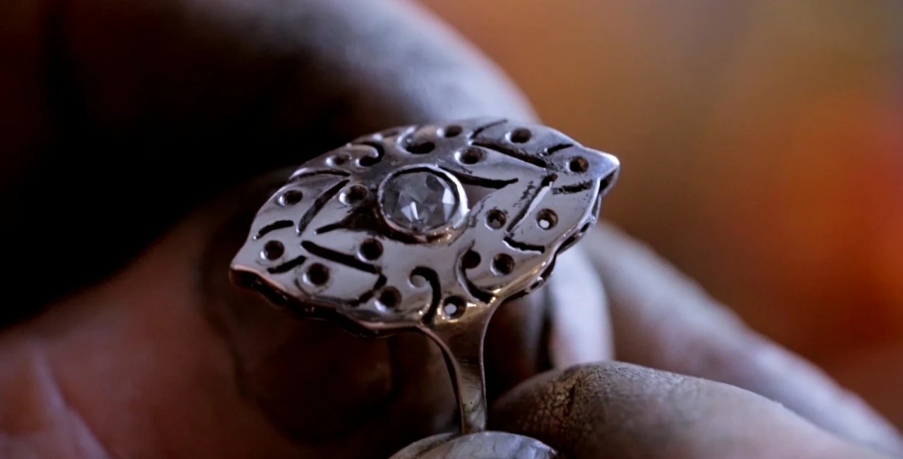 Biżuteria zaprojektowana i wykonana przez Jana Kalińskiego misternie wykonany srebrny pierścień z ozdobnym wzorem i osadzonym na środku kamieniem, trzymany przez dłoń o zabrudzonych palcach