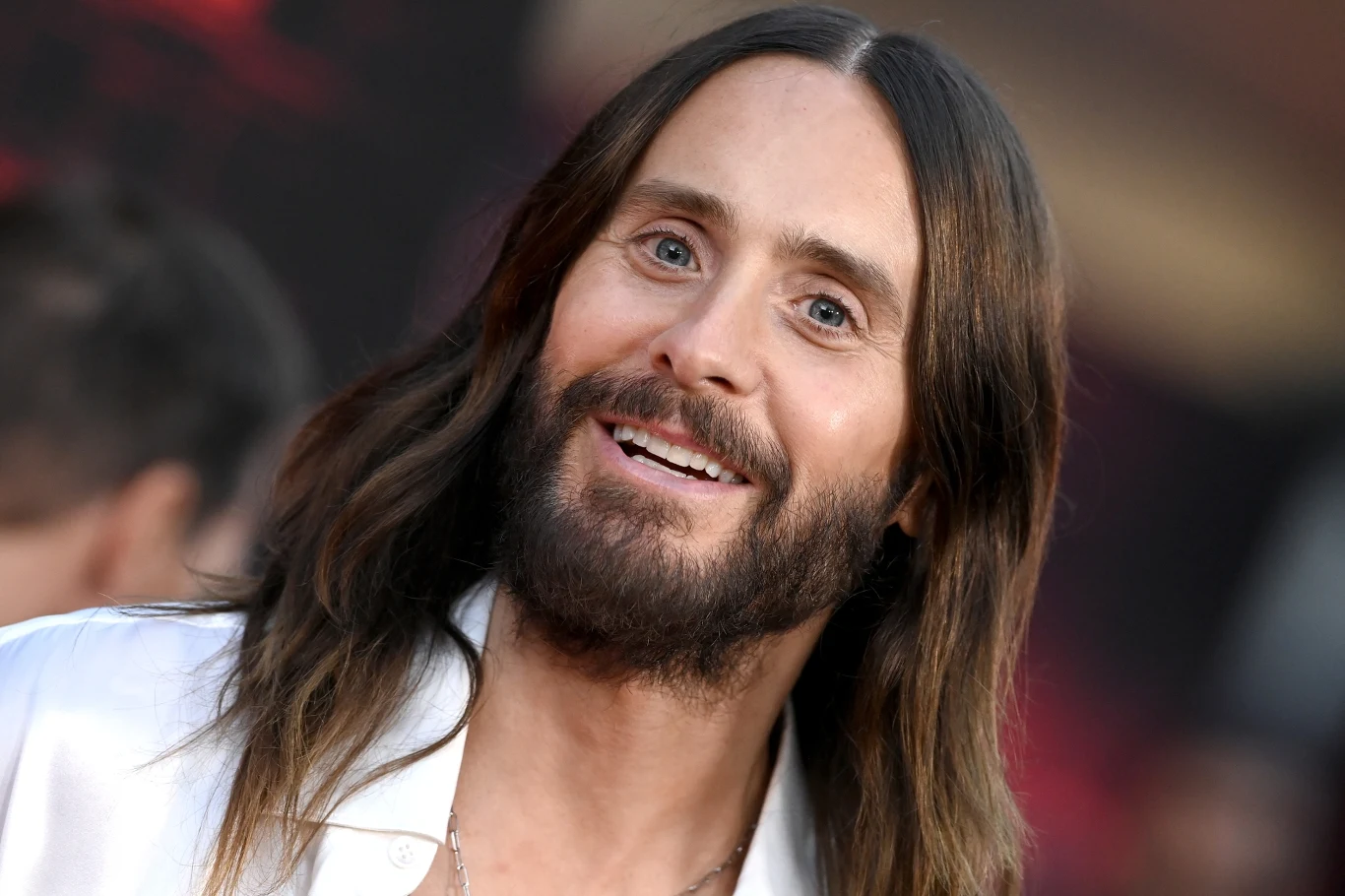 Jared Leto stoi na czele grupy Thirty Seconds to Mars Jared Leto, wokalista Thirty Seconds to Mars, z długimi brązowymi włosami i brodą, w białej koszuli na ciemnym tle.