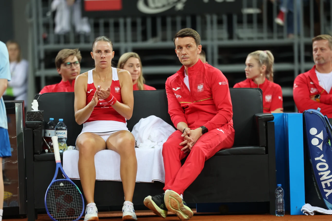 tenisistka w stroju reprezentacyjnym oraz trener w czerwonym dresie z orzełkiem siedzą razem na ławce przy korcie, obok leży rakieta i ręcznik, w tle widać członków ekipy oraz trybuny