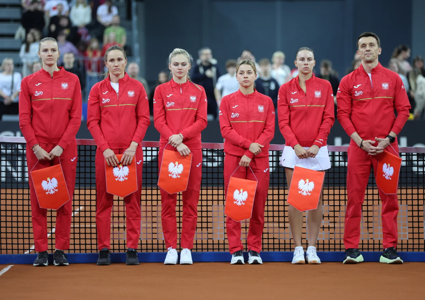 sześć osób w czerwonych dresach z godłem Polski stoi w równym szeregu na korcie tenisowym, trzymając flagi z białym orłem, w tle widać publiczność