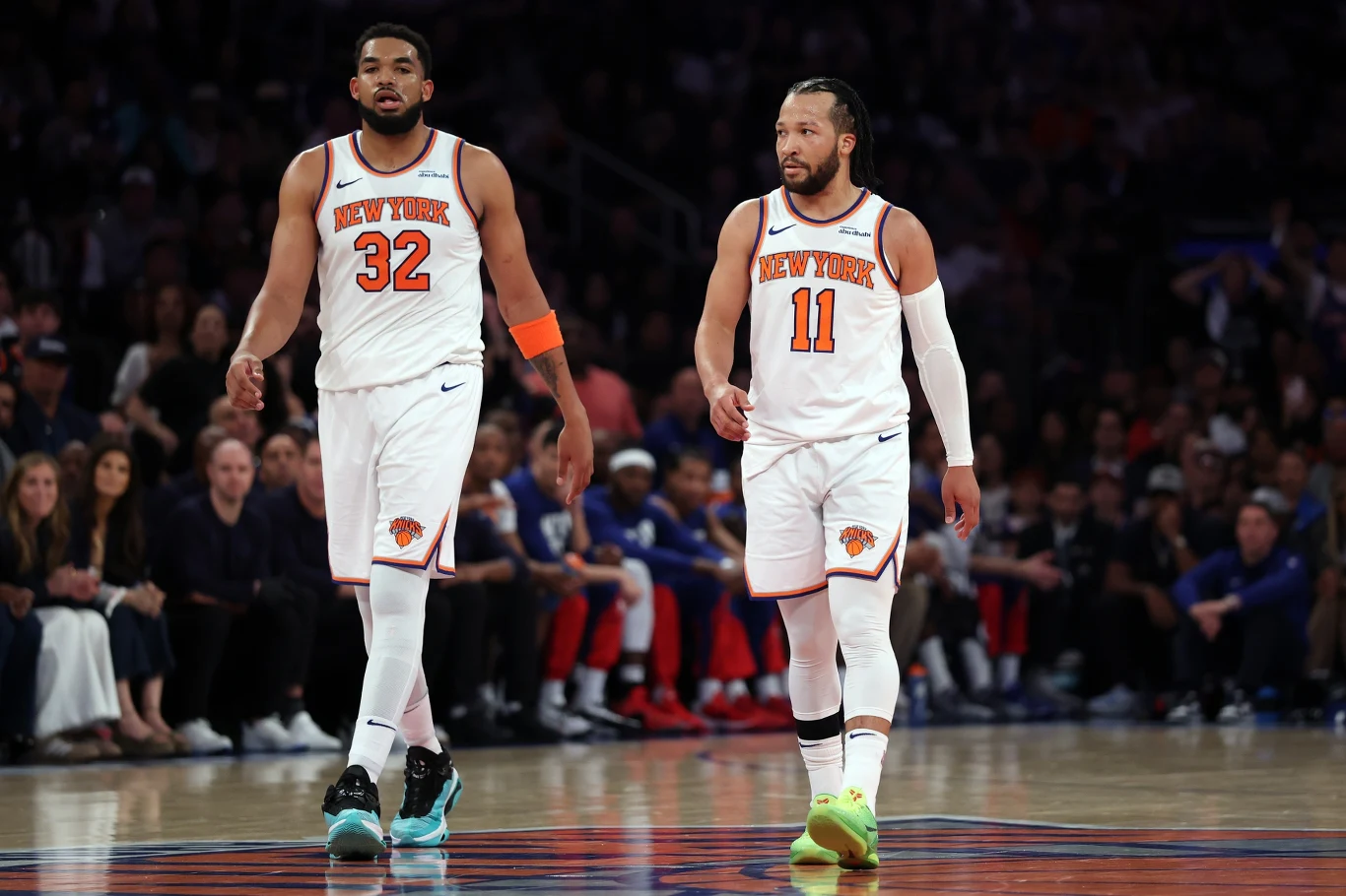Dwóch koszykarzy drużyny New York Knicks w białych strojach w trakcie meczu na parkiecie, otoczeni przez kibiców na trybunach; jeden zawodnik z numerem 32 nosi opaskę na ramieniu, drugi z numerem 11 zielone buty.